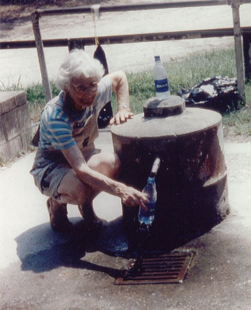 A woman kneels at the side of the spring, filling a plastic water bottle. Another bottle, already full, sits atop the spring.
