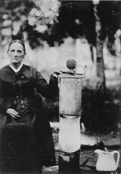 A elderly woman, in dark dress and with a dour expression, stands next to a wood post with a spout extending from the opposite side. Water flows from the spout into a pitcher on the ground. The top of the post sports a decorative wood ball, like a newell post.